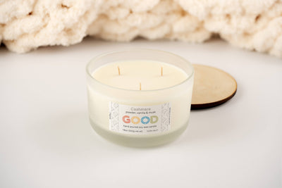 A white three-wick soy candle named "Cashmere" in a frosted glass jar sits on a white surface with its wooden lid beside it; a soft cream blanket appears in the background. The label notes powder, vanilla, and musk scents.