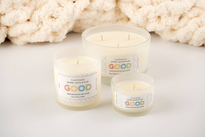 Four creamy white Cashmere soy wax candles in frosted glass jars of various sizes are displayed on a white surface, each with a GOOD label noting their hand-poured quality, set against a soft, textured fabric background.