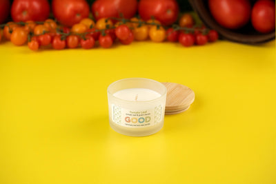 A small white Tomato Leaf soy wax candle rests on a bright yellow surface with its wooden lid nearby. Fresh tomatoes and cherry tomatoes in the background hint at the lush, garden-inspired scent of the Tomato Leaf candle.