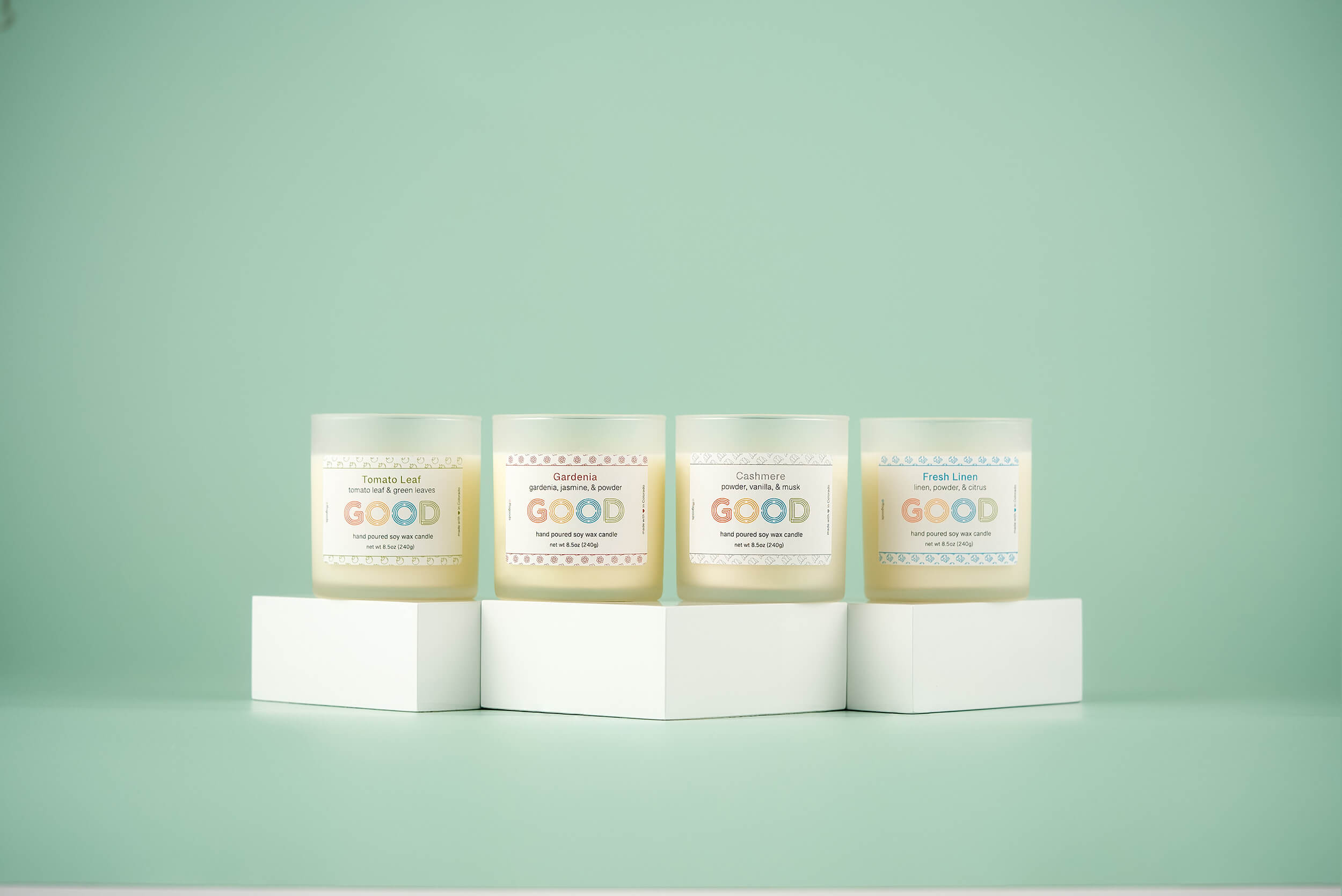 Four Frosted Stock candles in frosted glass jars are displayed on white blocks against a pastel green background. Each features a unique scent and colorful label—perfect for premium candle lovers, now available at a special discount.