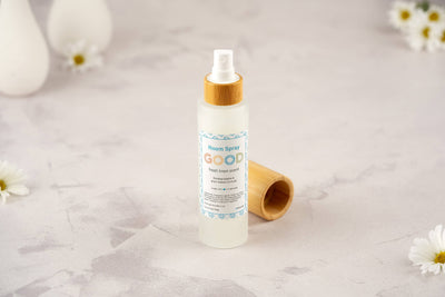 A clear spray bottle labeled “Room Spray” with a wooden cap sits on a light surface, framed by white daisies in the background—an ideal display for this plant-based Room Spray.