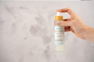 A hand holds a frosted glass spray bottle labeled Room Spray with a wooden cap, highlighting a plant-based air freshener against a softly blurred background.