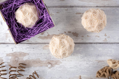Cream-colored Flower Orb Shaped Candle—crafted from beeswax soy, designed as clustered roses. One in a box with purple paper, two on a white wood surface with springs and burlap—an ideal Mother’s Day gift.