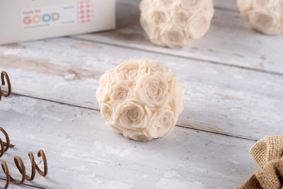 The Flower Orb Shaped Candle, a round ivory candle with intricate rose designs, rests on a rustic white wooden surface—an elegant Mother’s Day gift. A similar floral orb and blurred objects appear in the background.