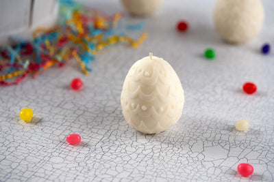 The Easter Egg Shaped Candle, with a vanilla scent, rests on a cracked white surface surrounded by colorful jellybeans and festive shredded paper.
