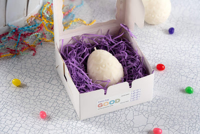 A white chocolate Easter egg in a white box with purple shredded paper is surrounded by colorful jelly beans, confetti, and the Easter Egg Shaped Candle on a textured white surface.