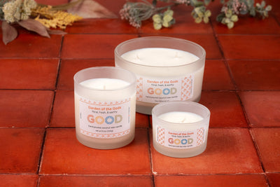 Three “Garden of the Gods” candles in frosted glass jars sit on a red tiled surface, evoking the sacred gardens fragrance of Colorado Springs, with dried flowers and greenery in the background.
