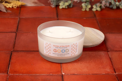 A white, three-wick candle labeled "Garden of the Gods" sits in a frosted glass container on red tiles, evoking Colorado Springs’ garden fragrance. The wooden lid is nearby, with decorative leaves and flowers in the background.