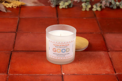 A white candle labeled Garden of the Gods rests in a frosted glass jar on red tiles, its wooden lid beside it. Inspired by Colorado Springs, it fills the air with sacred gardens fragrance as greenery and dried flowers blur in the background.