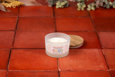 A white candle labeled Garden of the Gods sits in a frosted glass jar on terracotta tile, filling your space with its sacred gardens fragrance inspired by Colorado Springs. Dried flowers decorate the background.