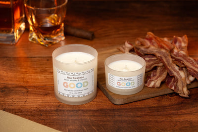 Two white Ron Swanson Candles, labeled "Good," sit on a wooden surface near a glass of whiskey and a board loaded with cooked bacon, creating a warm, cozy vibe.
