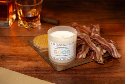 A limited edition Ron Swanson Candle sits on a wooden board beside cooked bacon strips, two whiskey glasses, and a candle lid on a rustic table.