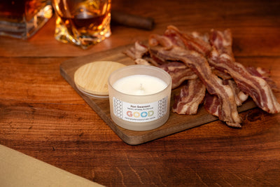 A lit Ron Swanson Candle in a glass jar labeled “GOOD” sits on a wooden surface beside its lid and a board with cooked bacon strips. Glasses of drinks and a cinnamon stick are visible in the background.