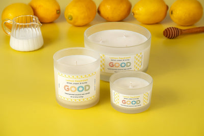 Three white Lemon Daydream candles in frosted glass jars sit on a yellow surface with fresh lemons, a jug of milk, and a honey dipper, creating an atmosphere of fresh lemon scent.