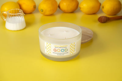 A white candle labeled Lemon Daydream sits in a frosted glass jar on a yellow surface with lemons, a small milk pitcher, and a honey dipper, creating a fresh lemon atmosphere.