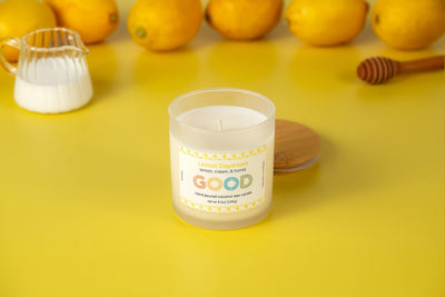 Lemon Daydream, a white lemon-scented candle in a refillable concrete vessel, rests on yellow with its wooden lid nearby. Surrounded by fresh lemons, a small pitcher, and honey dipper, it offers long-lasting fresh lemon fragrance.