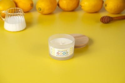 Lemon Daydream, a white candle with a fresh lemon scent in a refillable concrete vessel, rests on a yellow surface. Nearby are its wooden lid, fresh lemons, a honey dipper, and a small glass milk jug.