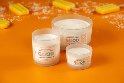 Three Sunshine Pop candles in frosted glass jars, labeled "Sunshine Pop," rest on an orange surface surrounded by yellow popsicles and white flakes, evoking their bright orange coconut scent.