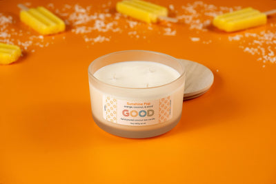The Sunshine Pop candle, featuring an orange coconut scent, sits in a frosted glass jar on an orange surface with yellow popsicles and coconut flakes nearby. Its wooden lid is placed beside the jar.