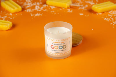 The Sunshine Pop candle, featuring an orange coconut scent, is displayed in a glass jar with its wooden lid beside it on an orange surface, surrounded by yellow popsicles and coconut flakes.