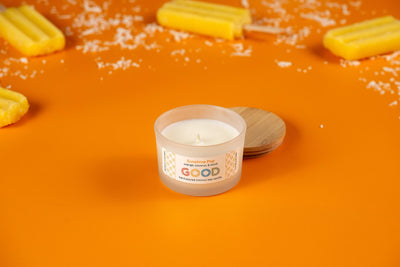 A Sunshine Pop candle in a frosted jar with wooden lid sits on an orange surface, surrounded by yellow popsicles and shavings. The label reads Sunshine Pop and features an uplifting orange coconut scent.