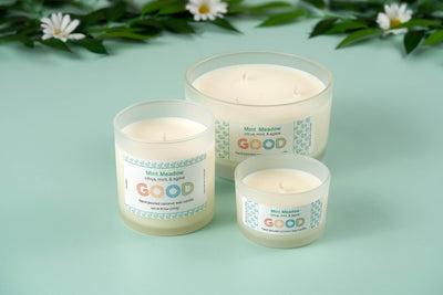 Three Mint Meadow candles in glass jars, each with a refreshing scent, are displayed on a light green surface. The jars, labeled "Mint Meadow," vary in size and are surrounded by green leaves and white flowers in the background.