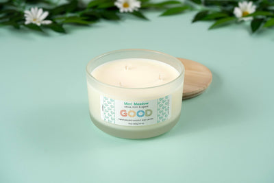 The Mint Meadow candle, in a frosted glass jar with a wooden lid, rests on a mint-green surface among white daisies and green leaves, capturing the refreshing essence of its signature scent.