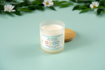 The Mint Meadow candle, in a frosted glass jar with a wooden lid, rests on a light teal surface. Its refreshing scent is highlighted by the soft blur of green leaves and white flowers in the background.