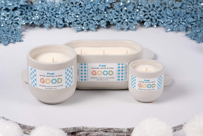 Three white Frost candles of varying sizes sit on a white surface, surrounded by artificial snow, blue snowflakes, and frosty branches for a winter-inspired display.