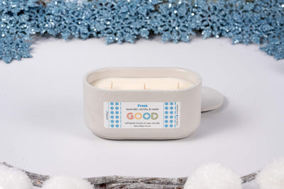 The Frost candle, hand-poured with lavender vanilla fragrance, sits in a white container on a snowy surface surrounded by blue snowflakes and faux snow, creating a cozy winter vibe.
