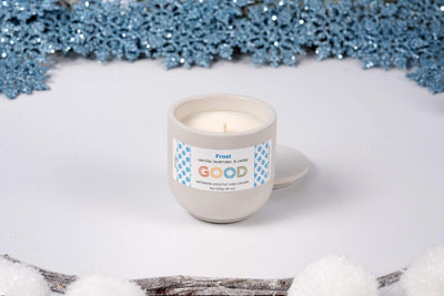 A hand-poured Frost candle in a round container, labeled “vanilla, lavender, & cedar. GOOD.” sits on a white surface with blue snowflakes and faux snow in the background.