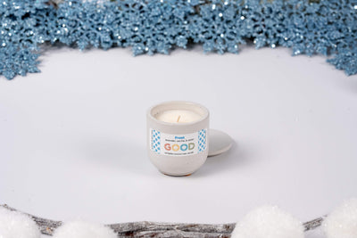 A hand-poured Frost candle in a small ceramic container sits on a white surface, surrounded by artificial snow, blue glittery snowflakes, and white decorations for a cozy winter vibe.
