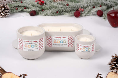 Three Cranberry Cheer candle jars sit on a white surface, surrounded by festive winter decorations, pinecones, apples, and cranberries, releasing a cheerful fruity and spiced aroma.
