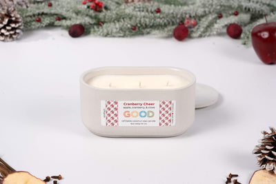A Cranberry Cheer candle in an oval white container rests on a white surface, surrounded by pinecones, apples, and festive greenery, capturing a holiday mood with its fruity-spiced scent.