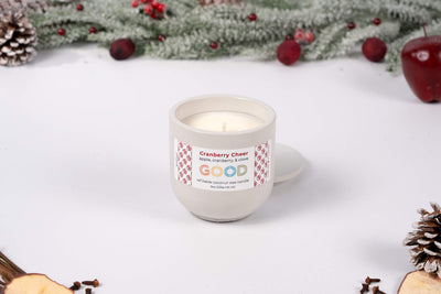 The Cranberry Cheer candle, in a white hand-poured concrete container, rests on a white surface with pinecones, greenery, red berries, and apple slices—evoking a festive fruity-spiced aroma for the season.