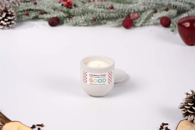 A Cranberry Cheer candle in a small concrete container sits on a white surface, surrounded by pinecones, frosted greenery, berries, and an apple to evoke its festive, fruity-spiced scent.
