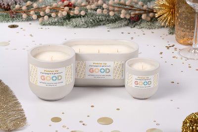 The Pinkies Up refillable candle, in elegant white and champagne hues, is displayed with festive gold decor, confetti, greenery, and a sparkling drink—making it the ideal centerpiece for any celebration.