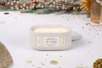 The Pinkies Up white oval champagne candle features three wicks and a “Pinkies Up, champagne & geranium, GOOD” label. Its hand-poured concrete vessel adds elegance to festive tables with gold accents, confetti, and greenery.