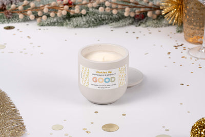 The Pinkies Up refillable scented candle in a round concrete container labeled "champagne & persimmon" sits on a festive white surface with gold confetti—an elegant choice for special occasions.