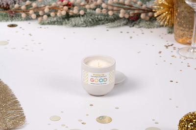 A Pinkies Up champagne-scented candle in a white ceramic holder labeled GOOD sits on a festive table with gold decorations, confetti, and greenery, creating a cheerful holiday atmosphere.