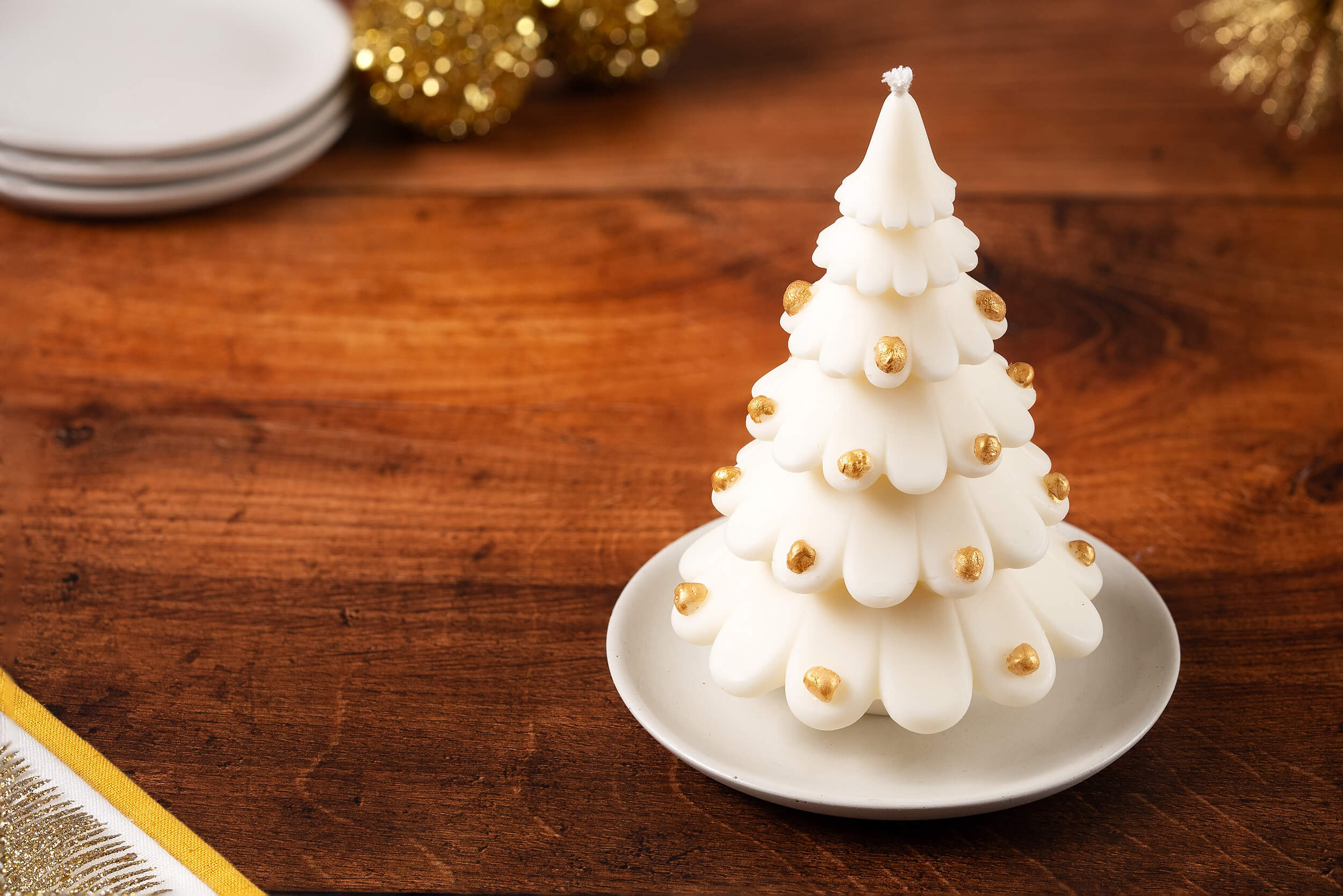 The Christmas Tree Shaped Candle, made from soy beeswax and adorned with gold beads, rests on a white plate on a wooden table with festive holiday decor in the background.