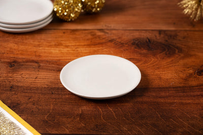 The Concrete Candle Plate, small and plain white, rests on a wooden table with stacked plates and golden holiday décor in the background.
