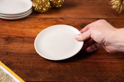 A hand holds a small Concrete Candle Plate over a wooden table, with stacked plates and gold decorative ornaments in the background.
