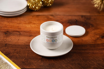 A white Pikes Peak Pine candle in a white jar sits on a Concrete Candle Plate with its lid beside it, displayed on a wooden table adorned with festive gold decorations and stacked plates in the background.