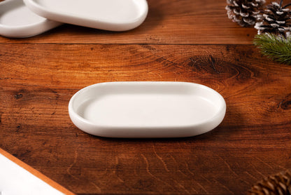 A small, white Concrete Candle Tray rests on a wooden surface with two stacked trays behind it and pinecones and evergreen branches in the background—ideal as a handcrafted, minimalist decor accessory for cozy spaces.