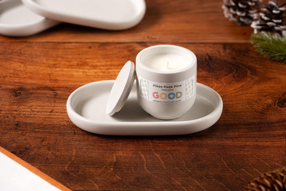 A white candle labeled Pikes Peak Pine with a partially open lid sits on a Concrete Candle Tray atop a wooden surface. Pinecones and greenery blur in the background, enhancing the minimalist home decor aesthetic.