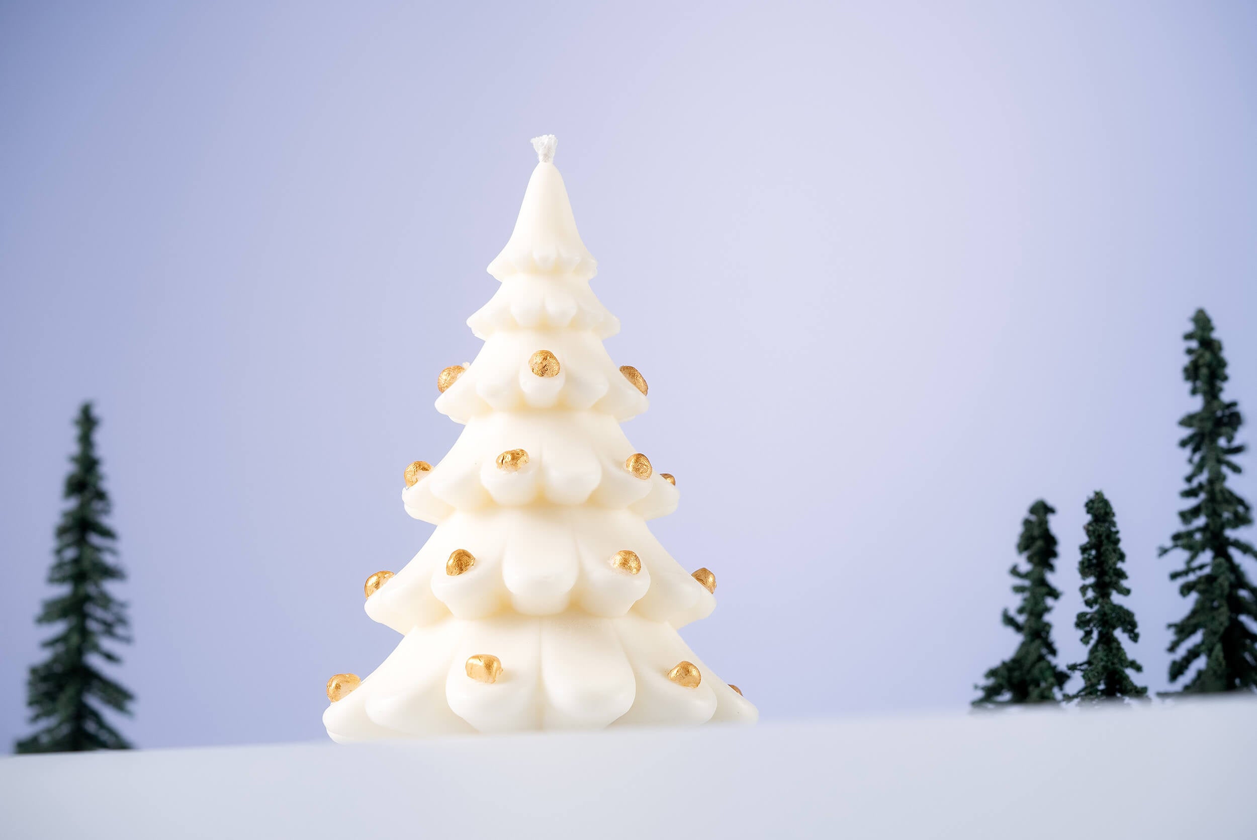 The Christmas Tree Shaped Candle, crafted from soy beeswax and adorned with small gold ornaments, stands on a snowy surface with miniature evergreens in the background—a perfect holiday decor accent.