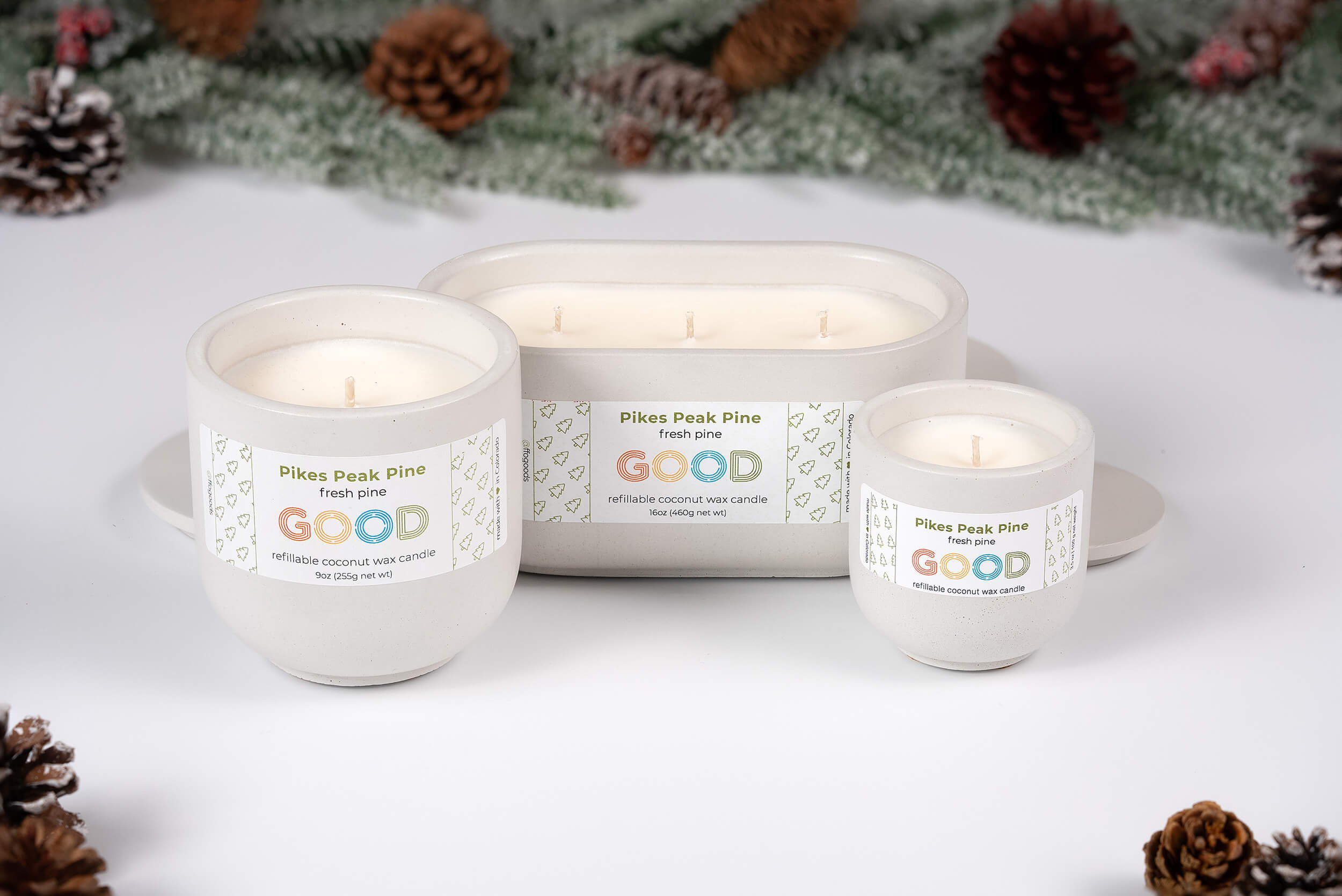 Three Pikes Peak Pine candles in varying sizes are arranged with pinecones and evergreens on a white surface, capturing a festive winter vibe—ideal for those who love the scent of Colorado pine.