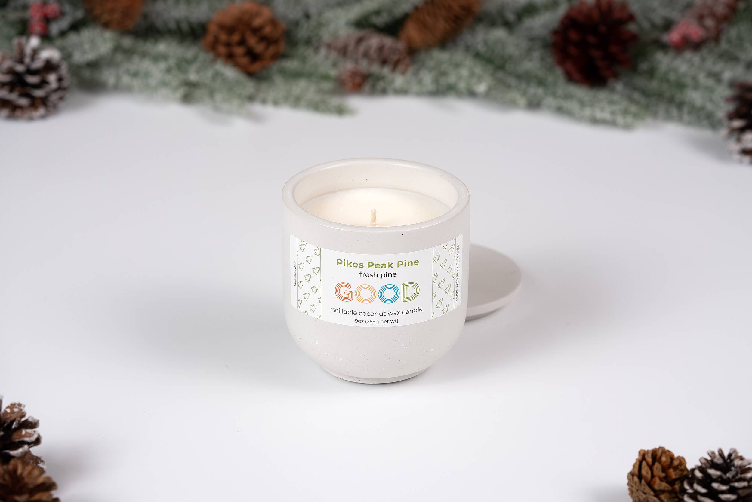A Pikes Peak Pine candle in a glass jar sits on a white surface, surrounded by pinecones and frosted pine branches. The lid is beside the candle, evoking the fresh scent of Colorado’s forests.