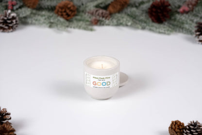 A Pikes Peak Pine candle in a small white ceramic container rests on a white surface, surrounded by pinecones and green pine branches in the background.
